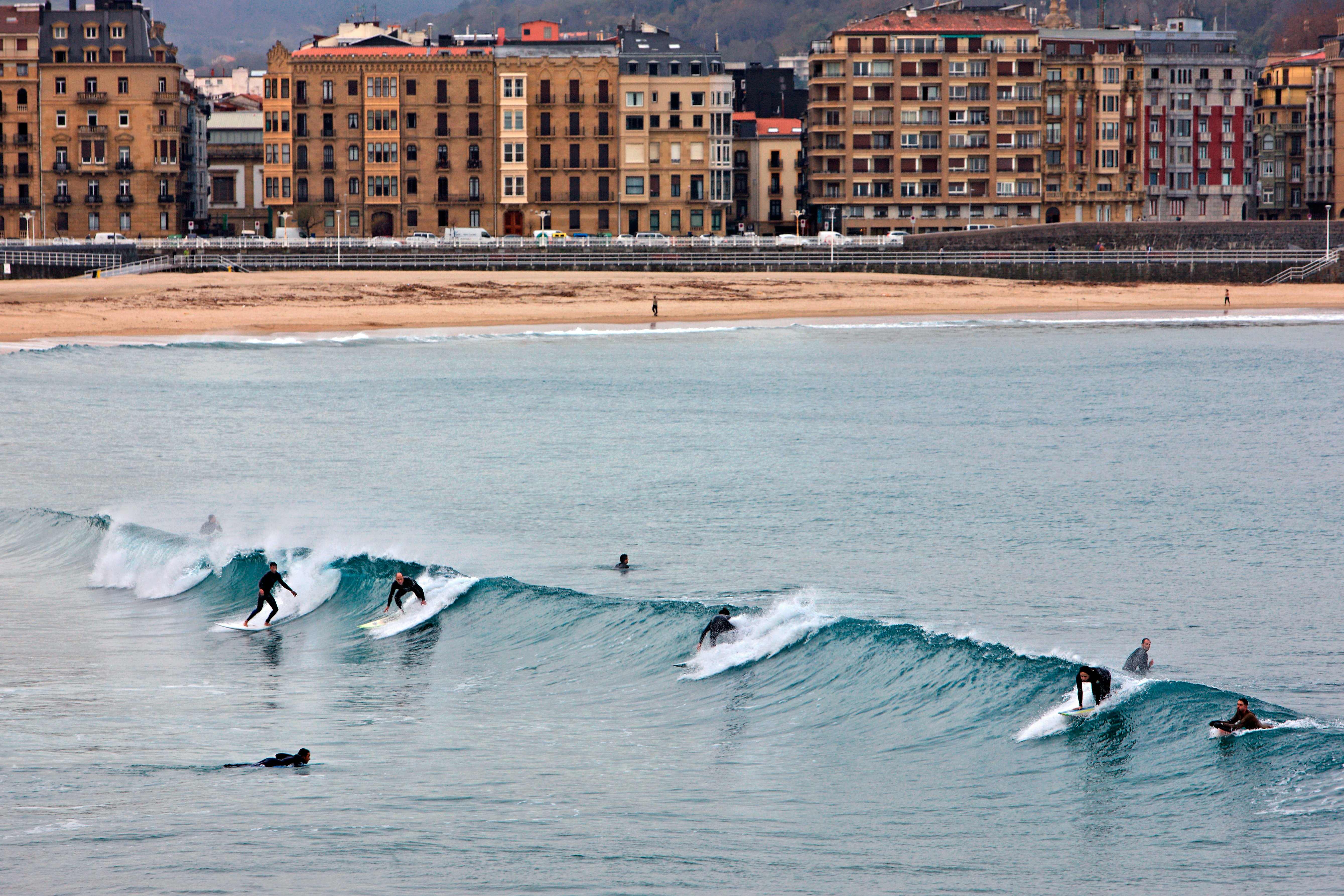 Surfers in wetusits catch a wave in the ocean near a city beach.